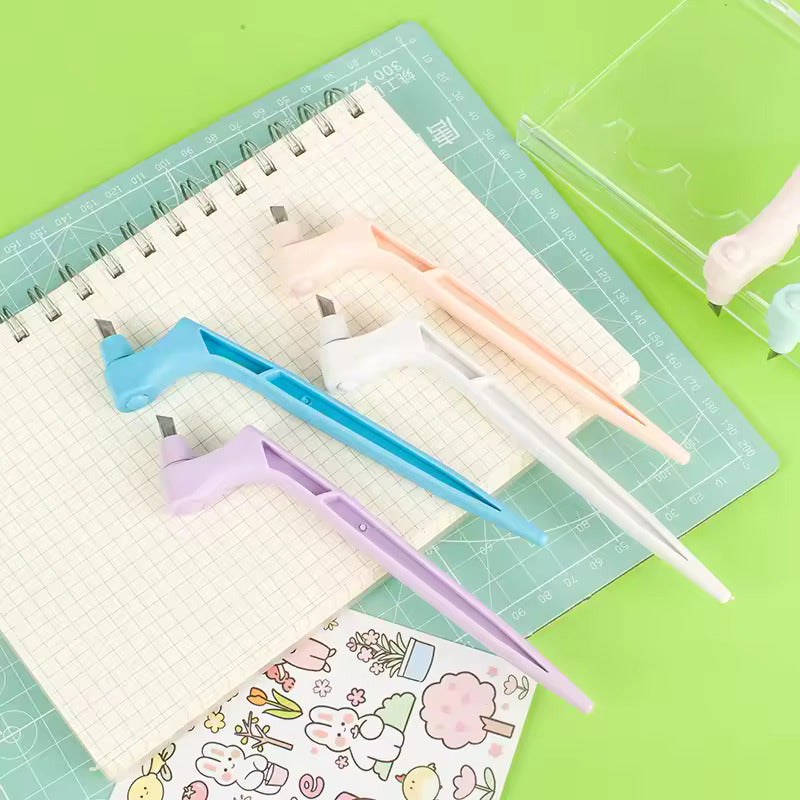Six colorful utility knives with 360-degree rotating blades are showcased on a grid-patterned notepad. Accompanied by a clear case and decorative stickers against a green backdrop, these "360 Degree Rotating Blade 6 Pcs" tools enhance both style and precision for your DIY projects.