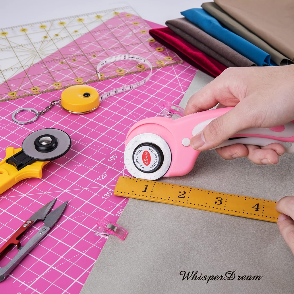 A person uses the 39-Piece Quilting Kit: Perfect for Beginners—including a rotary cutter and acrylic ruler—to cut fabric on a pink self-healing mat, surrounded by sewing tools and folded fabric.