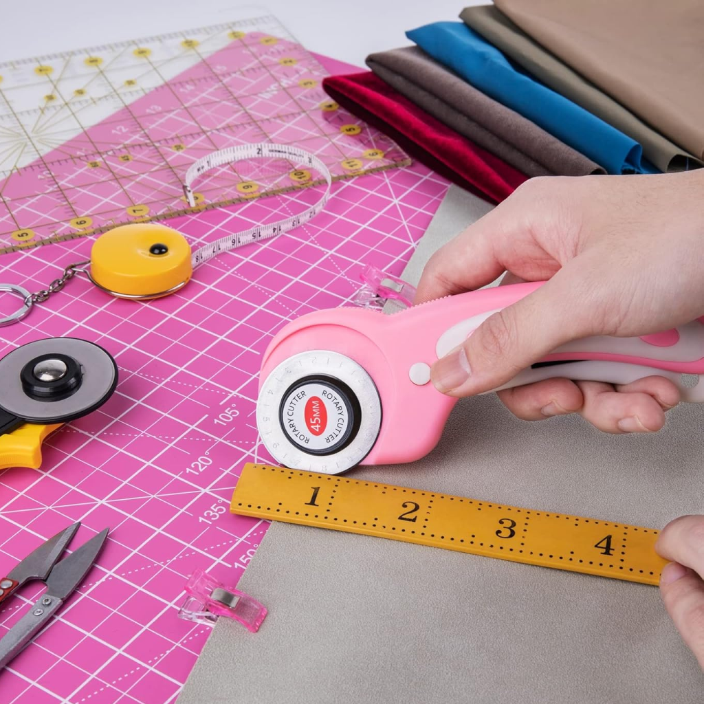 A person uses the 45mm Rotary Cutter with 5 Extra Blades and a ruler to cut fabric on a pink cutting mat, surrounded by sewing tools and measuring accessories.