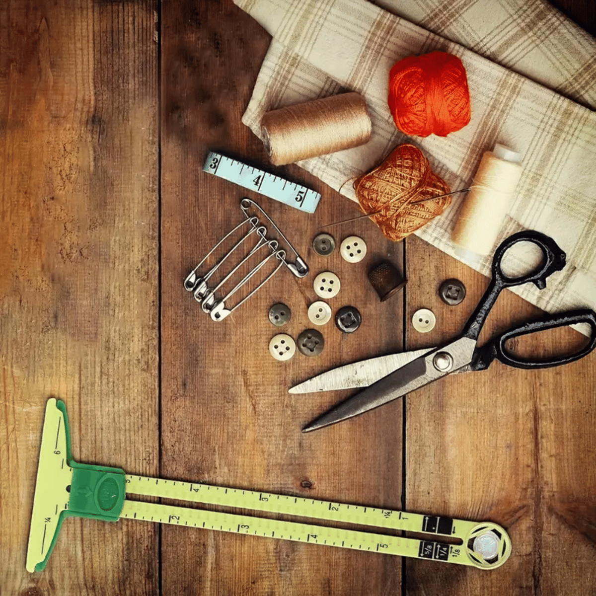 The Sliding Gauge Ruler is arranged with scissors, safety pins, thread spools, buttons, and fabric—perfect for precision measurements in quilting projects—displayed neatly on a wooden surface.