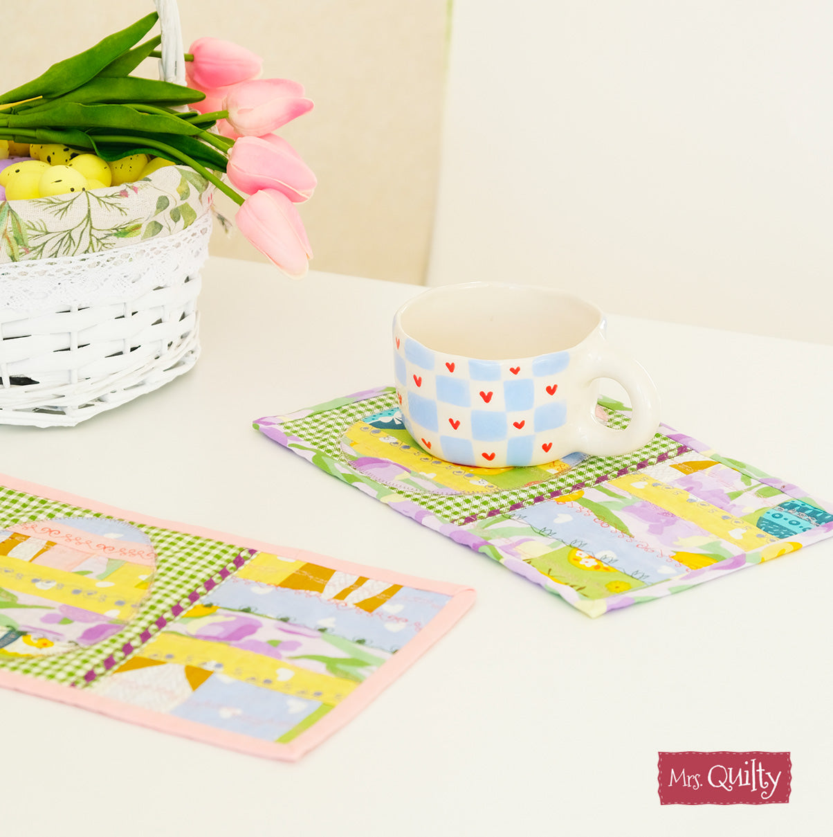 A white mug adorned with blue and red heart designs rests on a quilted coaster. Beside it, a white basket filled with pink tulips and vibrant eggs enhances the festive decor, beautifully complemented by the patchwork quilt which can be crafted using patterns from the Spring Magazine PDF download.