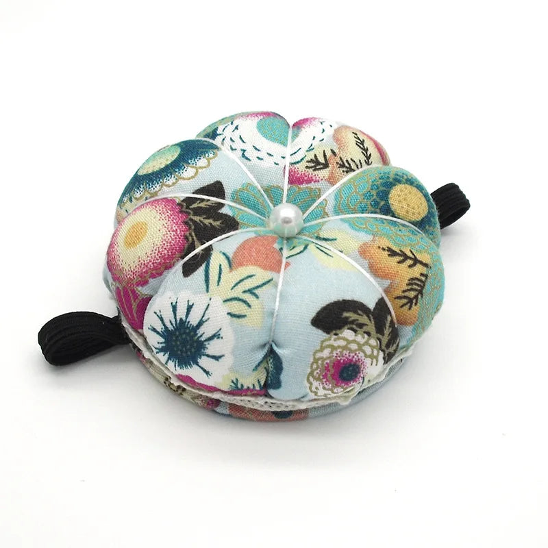 The Pin Cushion is a round fabric accessory with floral patterns, a central pearl-like bead, and black elastic bands. It's perfect for organizing sewing pins while you work on projects at any sewing station.