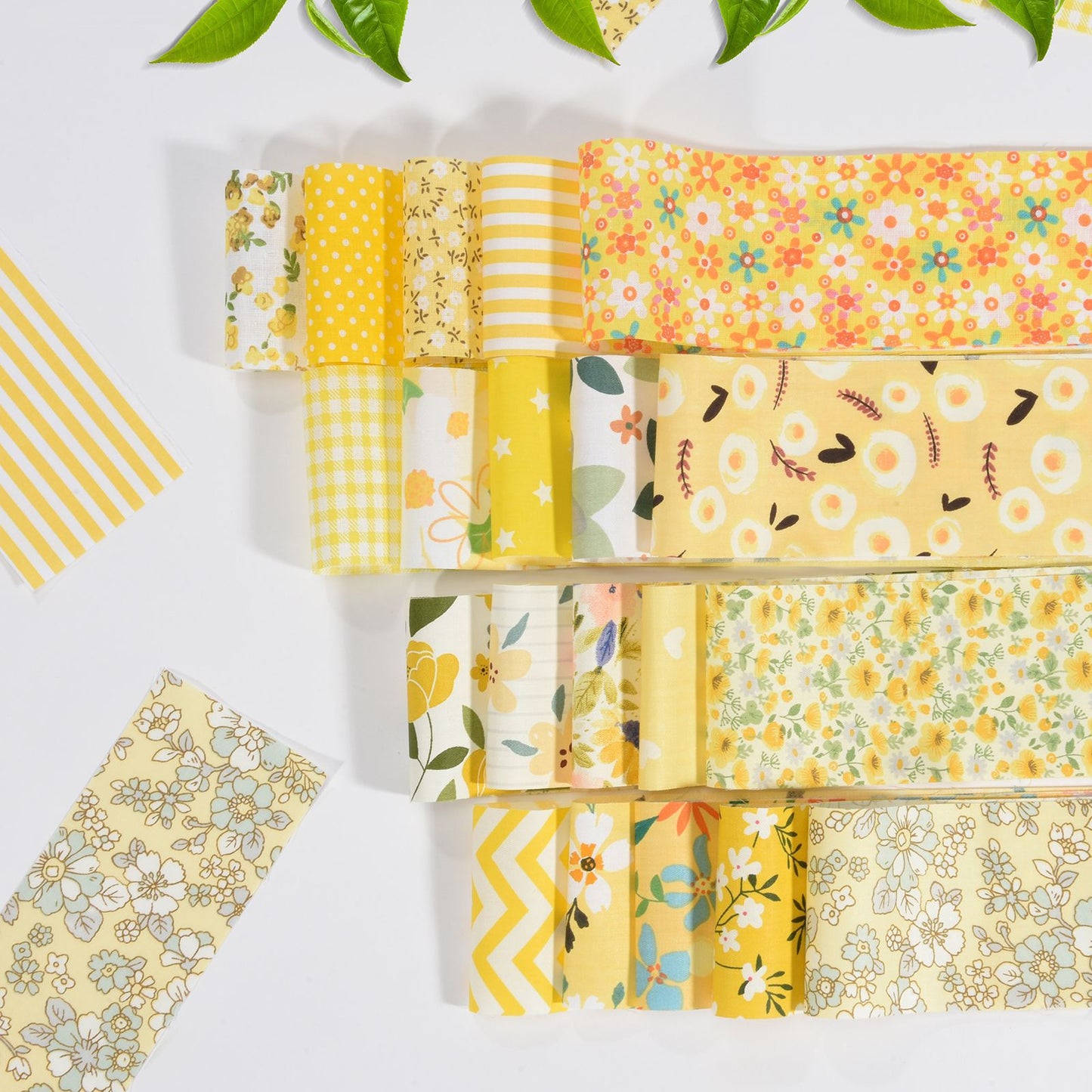 The Sunny Bloom Jelly Roll Bundle features assorted yellow fabric strips with floral patterns, beautifully arranged on a white surface and accented by green leaves—perfect for your next floral quilt project.