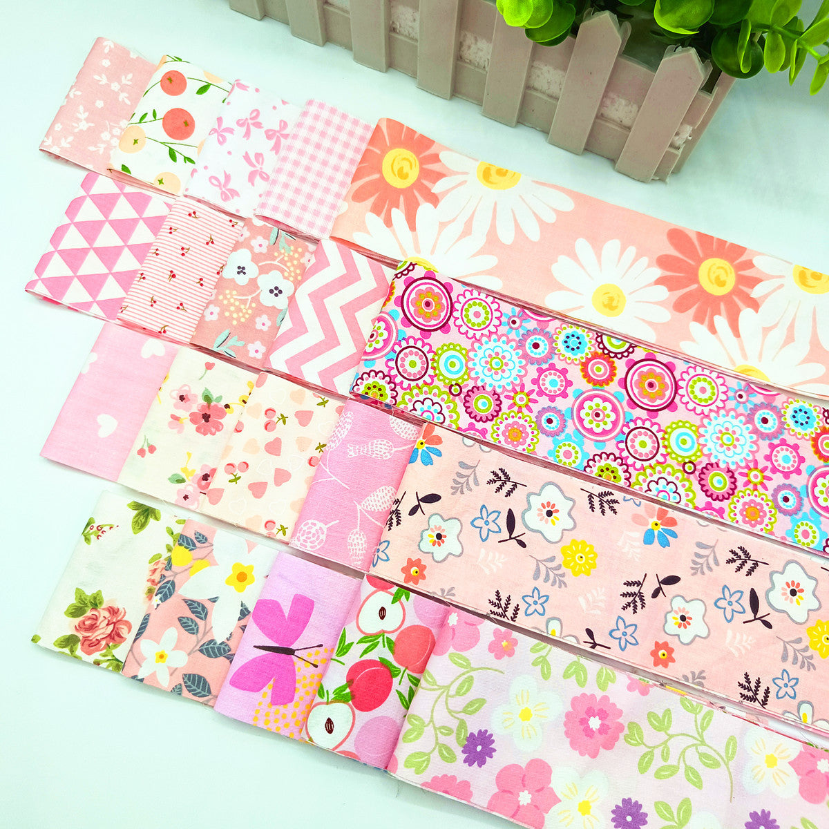 Rows of rectangular Pink Blossom Jelly Roll Bundle fabric pieces are arranged on a white surface, with a decorative green plant in the background.