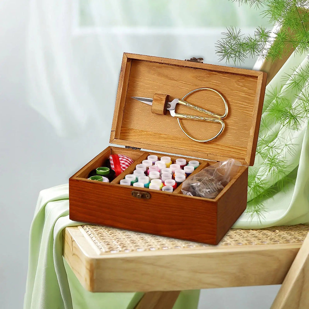 The Wooden Sewing Box with Lid epitomizes organized sewing storage, open on a chair to reveal neatly arranged scissors, thread spools, and buttons against a lush green cloth and foliage backdrop displaying essential sewing tools.
