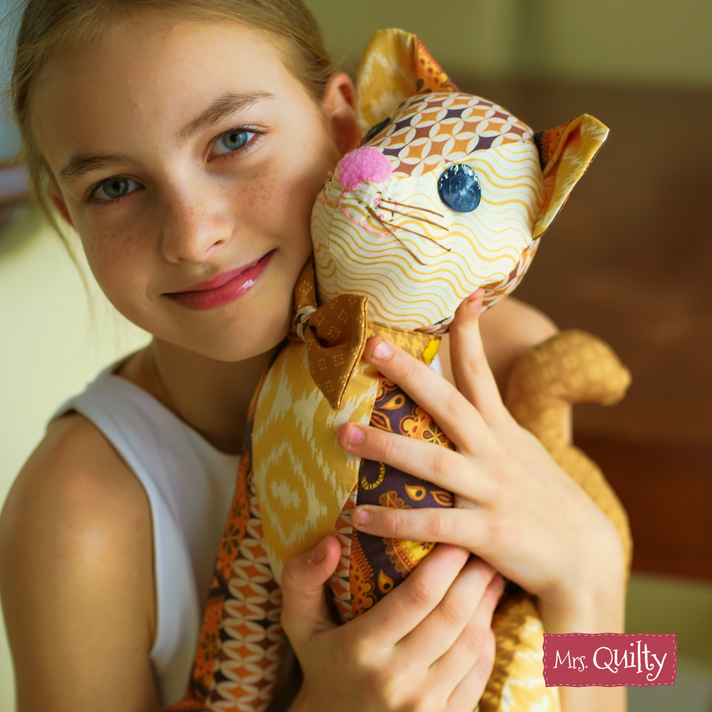 A young girl smiles while holding a plush quilted cat made from the "Callum the Cat Downloadable PDF Quilt Pattern," making it a perfect handmade gift.