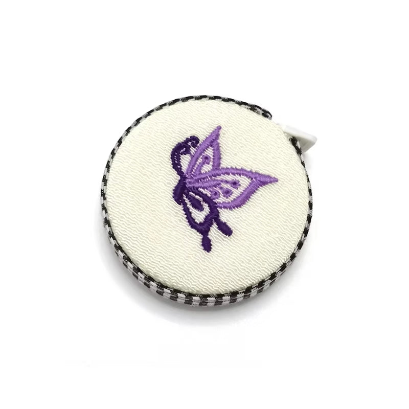The Retractable Fabric Measuring Tape 59inch (150cm) features a black and white border with a purple butterfly design on cream, making it both an elegant addition to your collection and a functional sewing tool.