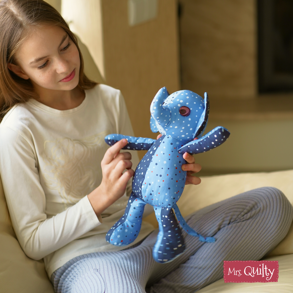 A girl sits on a couch holding an Ellie the Elephant toy made from blue polka dot fabric using the Ellie the Elephant Downloadable PDF Quilt Pattern. The Mrs. Quilty logo is visible in the bottom right corner.