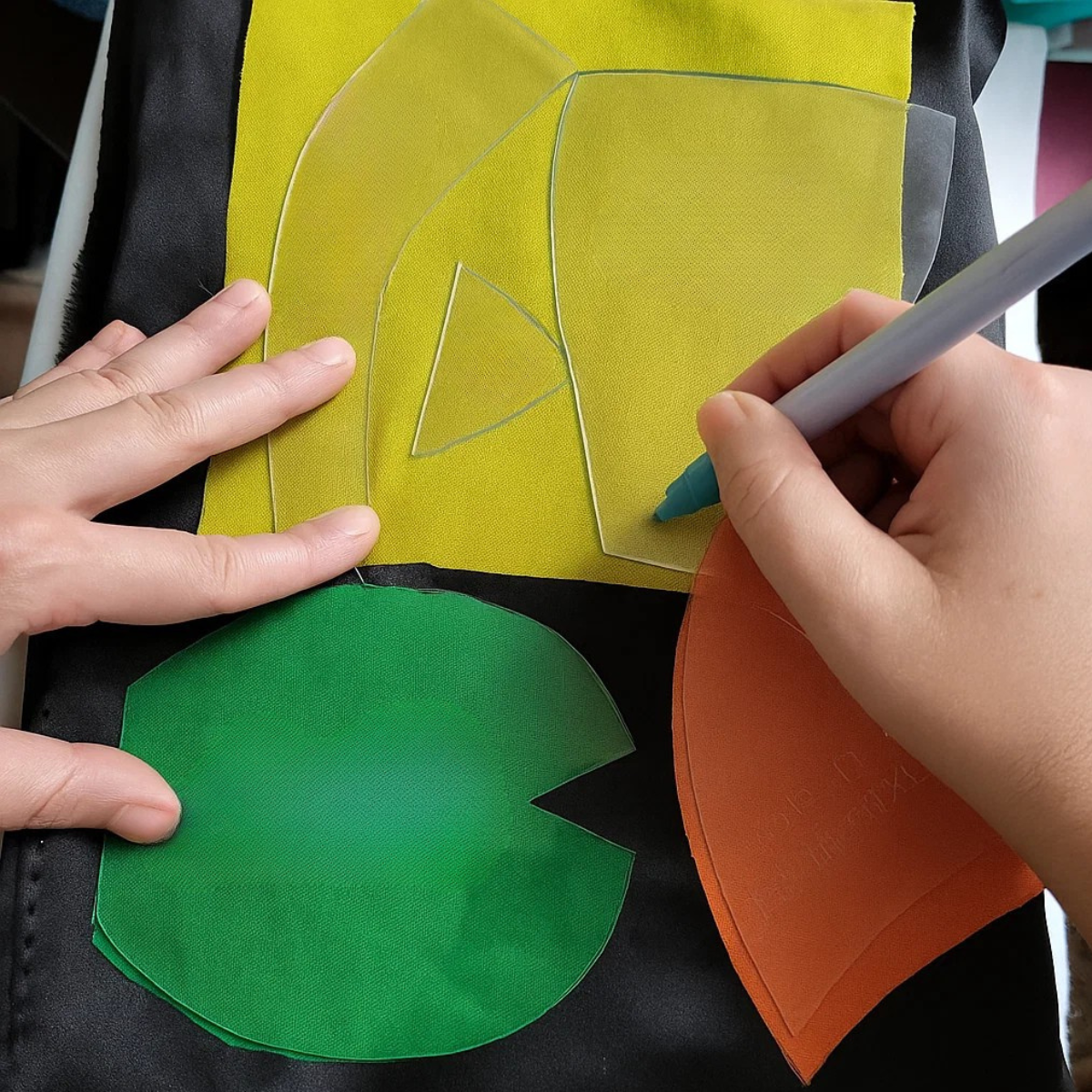 Two hands use the Acrylic Fox Doll Template Set to trace shapes onto yellow, green, and orange fabric with a pen on a black surface, starting a creative fox plushie DIY project.