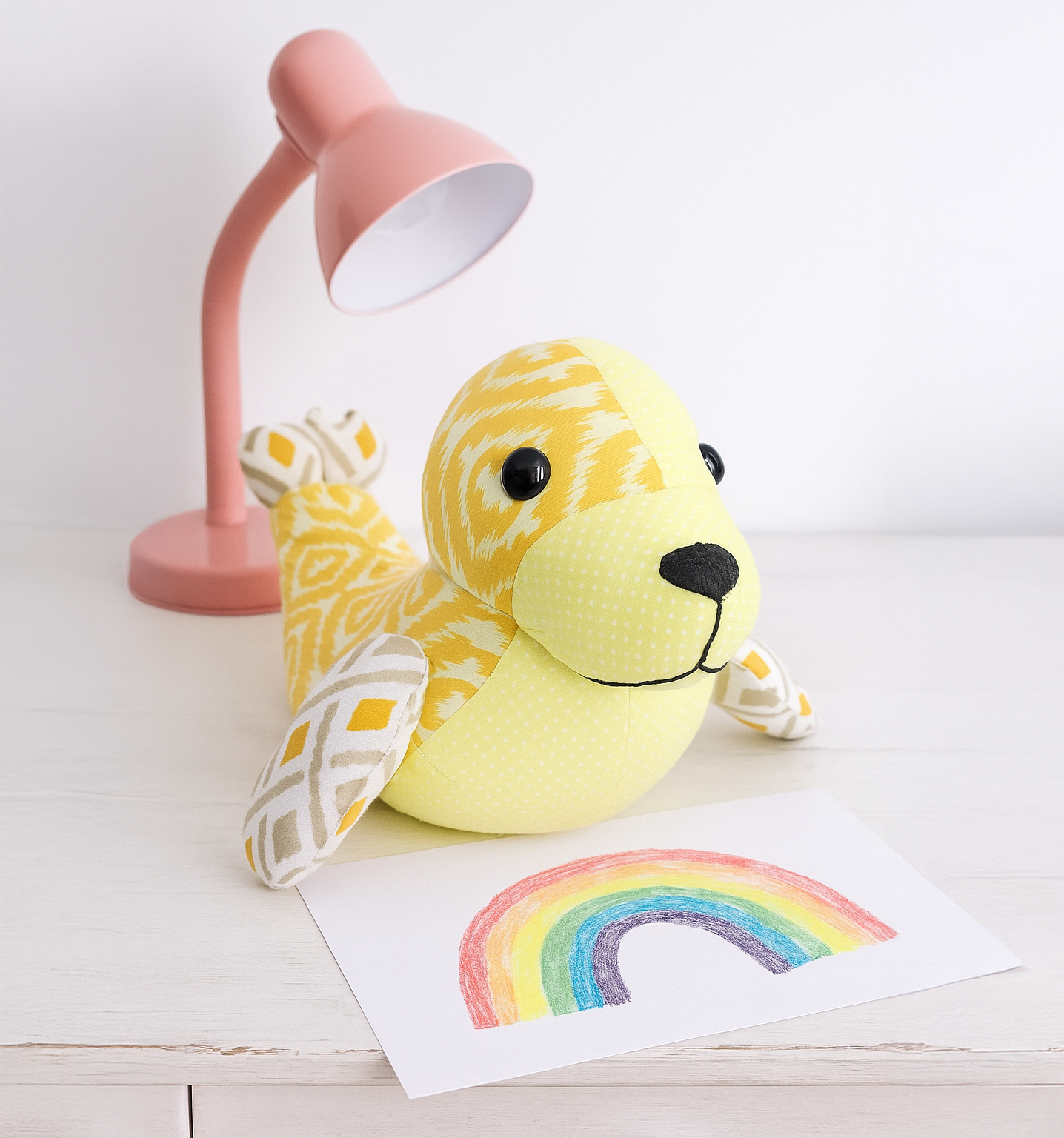 Tidey the Seal, made from the downloadable PDF quilt pattern, sits on a white table beside a pink desk lamp and a crayon rainbow drawing.