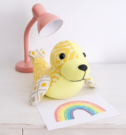 Tidey the Seal, made from the downloadable PDF quilt pattern, sits on a white table beside a pink desk lamp and a crayon rainbow drawing.