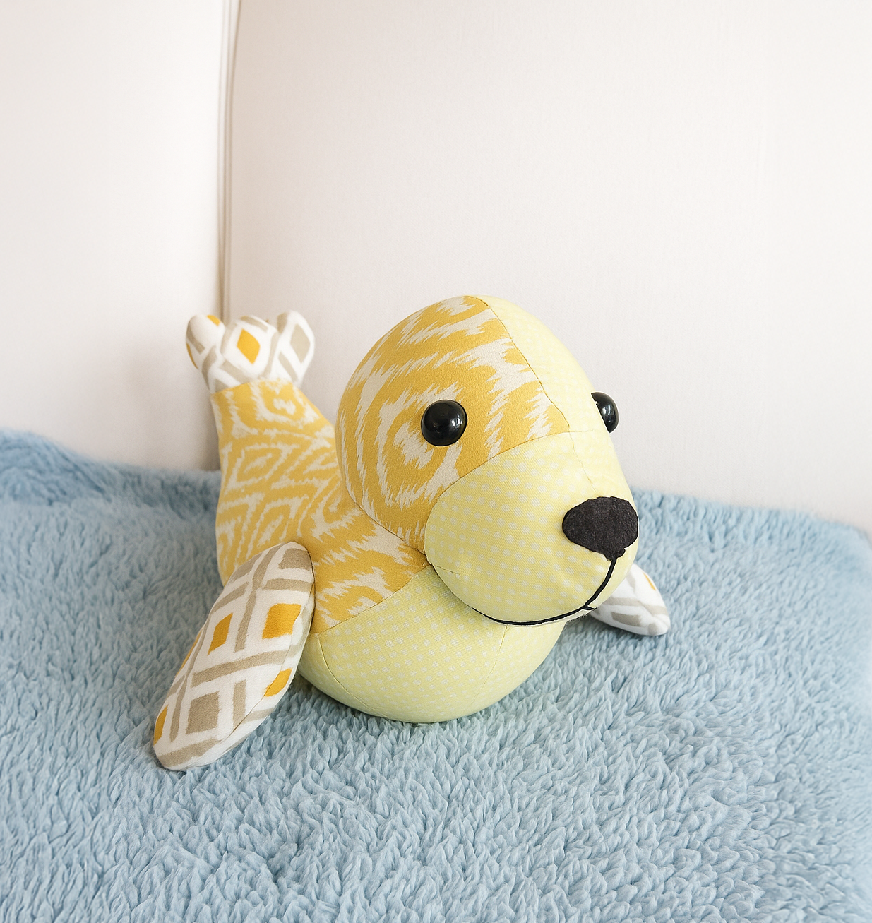 “Tidey the Seal Downloadable PDF Quilt Pattern features a yellow and white patterned ocean animal design displayed on a light blue textured blanket against a white background.”.