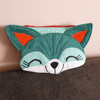 The Freddie Fox Purse Downloadable PDF Quilt Pattern lets you create a whimsical fabric pouch shaped like a fox's face, featuring teal and orange ears, a red zipper, and black embroidered features.