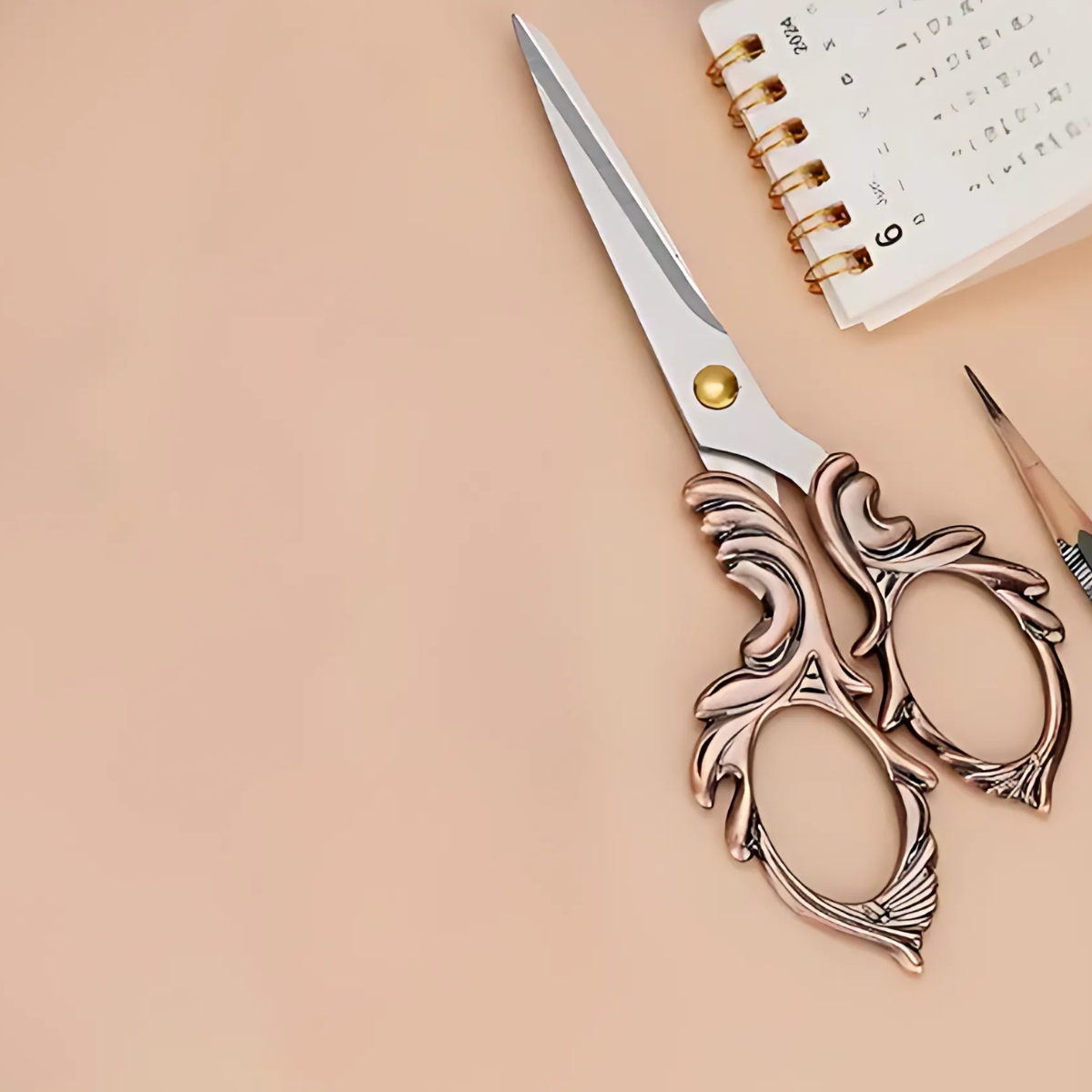 Embroidery Scissors, a pencil, and a small spiral-bound calendar are arranged on a beige surface, making these tools ideal quilting and crafting essentials.