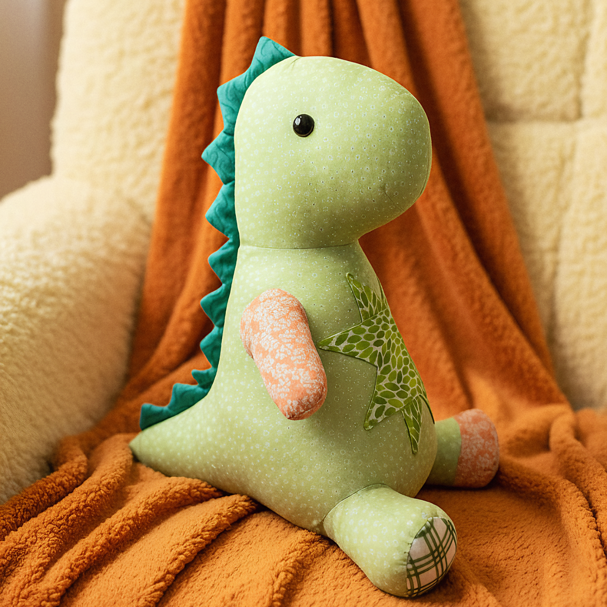 A light-colored chair is draped with an orange blanket featuring the playful "Rexie the Dino Downloadable PDF Quilt Pattern," evoking a green dinosaur toy accented with orange and plaid details.