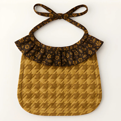 The Ruffled Bib Downloadable PDF Quilt Pattern features a brown floral collar and geometric houndstooth design—great inspiration for beginner quilting projects. The bib ties at the neck.