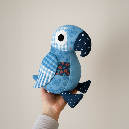 A hand holds Pip the Parrot, a stuffed toy featuring a colorful patchwork quilt design with stripes, flowers, and polka dots, displayed against a plain light background. The pattern is available as a downloadable PDF.