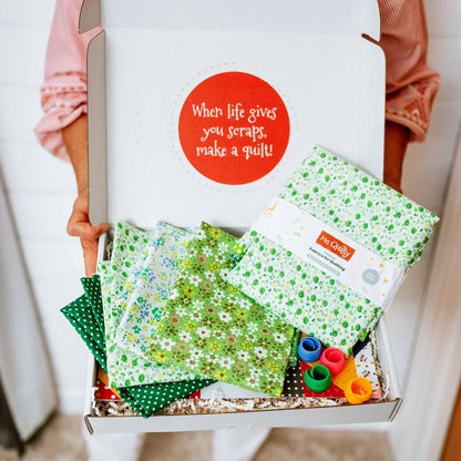 Open your [12-Month Prepaid] Mrs Quilty Subscription Box filled with green floral fabrics, thread spools, and a sign reading "When life gives you scraps, make a quilt!.