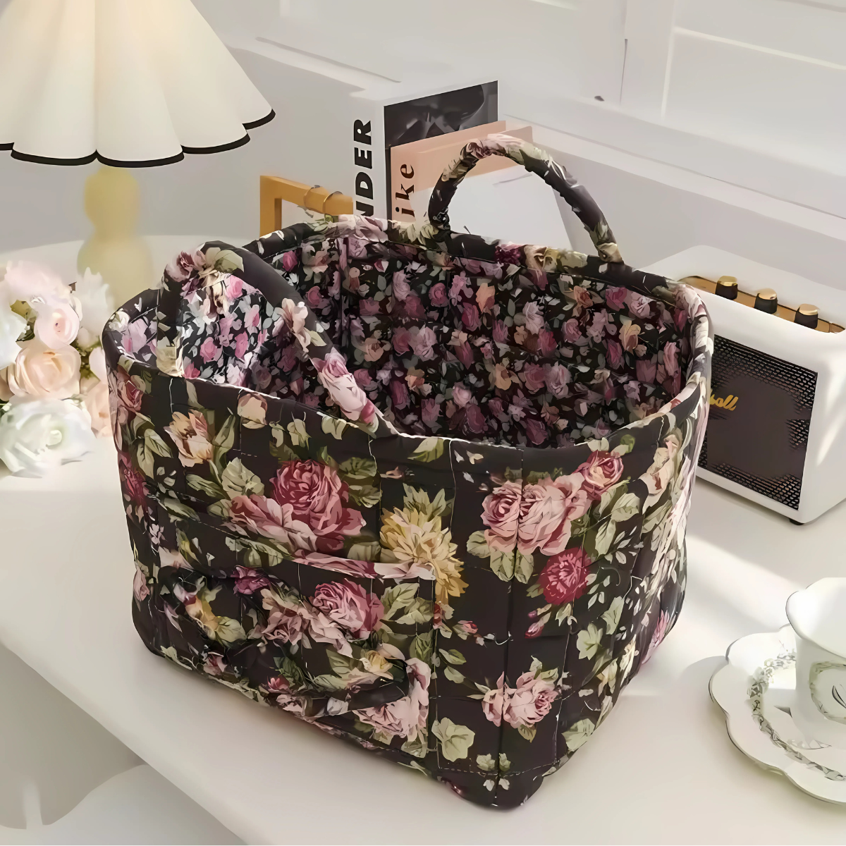 The Quilted Storage Basket, made from pure cotton with a floral design, rests on a white desk beside a lamp, books, flowers, a speaker, and plates—bringing vintage charm to your space.