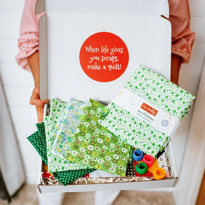A person holds an open [3-Month Prepaid] Mrs Quilty Subscription Box filled with green floral fabrics and colorful thread reels, featuring a quote: "When life gives you scraps, make a quilt!.