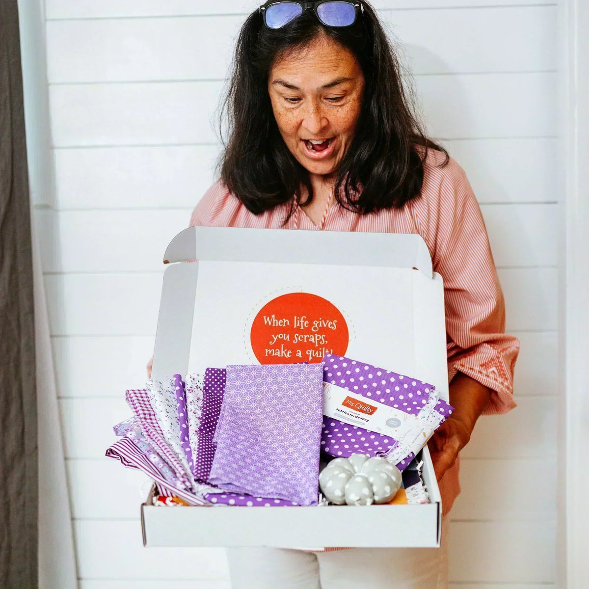 A person in a striped shirt holds an open [12-Month Prepaid] Mrs Quilty Subscription Box containing purple and white fabric pieces and round objects, with a motivational message on the inside lid.