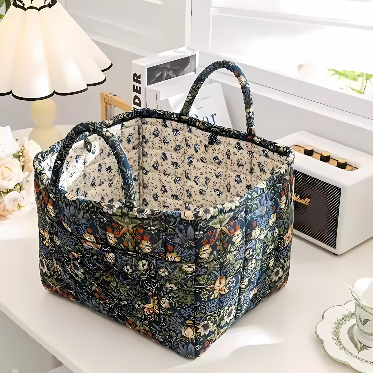 The Quilted Storage Basket, made from pure cotton with a floral design, sits on a white desk near a lamp, books, and speaker, adding vintage charm to the bright room.