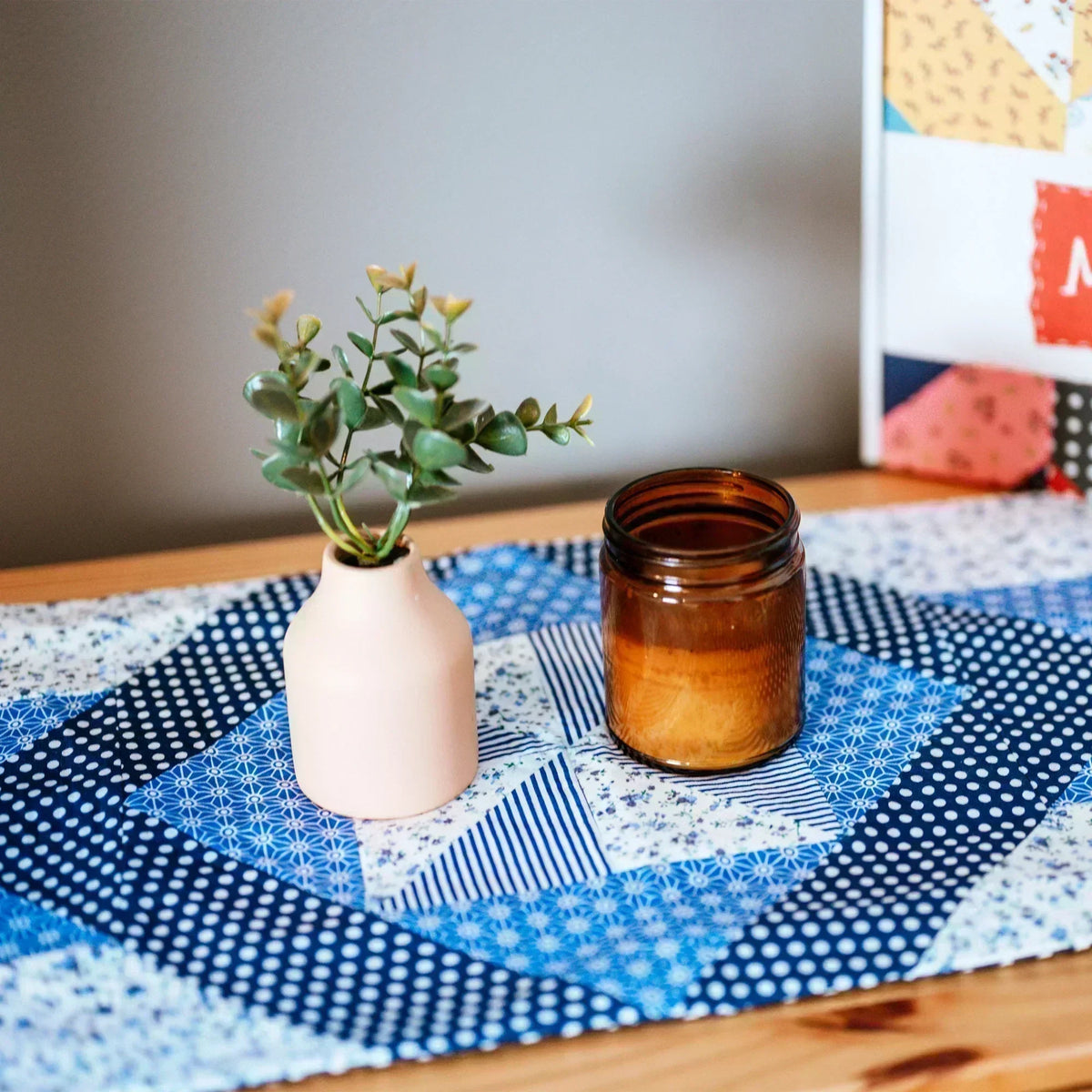 A [12-Month Prepaid] Mrs Quilty Subscription Box featuring a beige vase with a green plant and brown candle jar on a wooden table with blue geometric-patterned cloth.