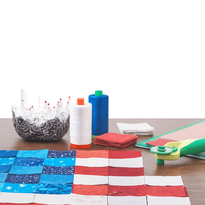 A table with July 4th Patterns Downloadable PDF Quilt Patterns, spools of thread, red, white, and blue fabric squares for Independence Day quilting, pins in a bowl, a rotary cutter, and a cutting mat.