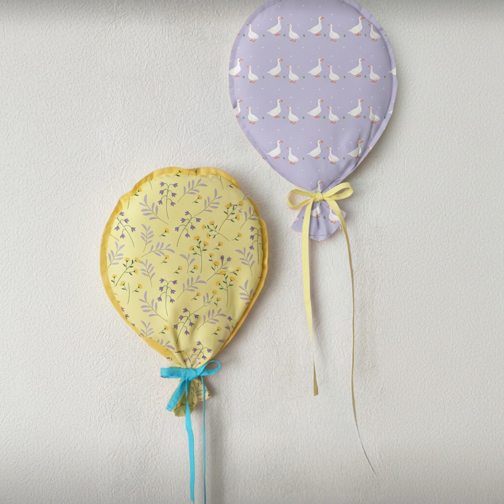 The Balloon Wall Hanging Downloadable PDF Quilt Pattern features two fabric balloons—one yellow floral, one lavender with geese—each with ribbon tails, creating a whimsical decor piece perfect for mounting on a white textured wall.