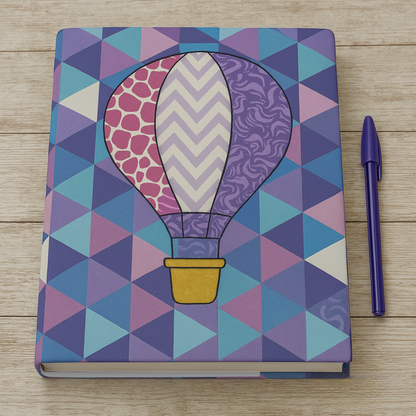 A downloadable PDF quilt pattern for a book cover featuring a vibrant geometric hot air balloon design, displayed with a blue pen on a wooden surface.