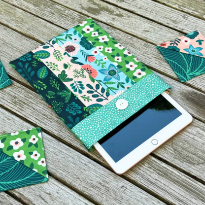 A tablet in a fabric sleeve made with the Book Sleeve Downloadable PDF Quilt Pattern featuring floral and leaf quilting rests on wood, alongside matching coasters.