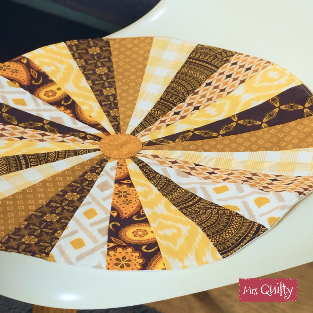 A downloadable PDF quilt pattern for a round, quilted patchwork chair cushion in brown, yellow, and white pinwheel designs is shown on a white chair. The “Mrs. Quilty” logo appears in the corner.