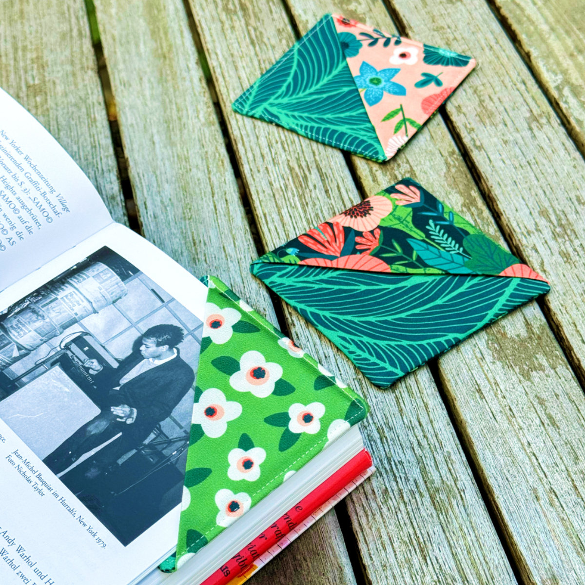 Three colorful corner bookmarks made with floral fabric scraps, including one in use, rest on a wooden surface—a delightful project using the Corner Bookmark Downloadable PDF Quilt Pattern for any book lover.