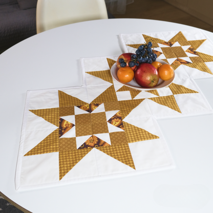 A white round table with a star-patterned quilted table runner made from the Table Runner Downloadable PDF Quilt Pattern, and a bowl of oranges, apples, grapes, and a plum.