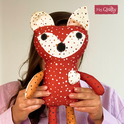 A person in a pink shirt holds a red and orange fox plushie with embroidered details, inspired by the "Finley the Fox Downloadable PDF Quilt Pattern." "Mrs. Quilty" appears in the top right corner.