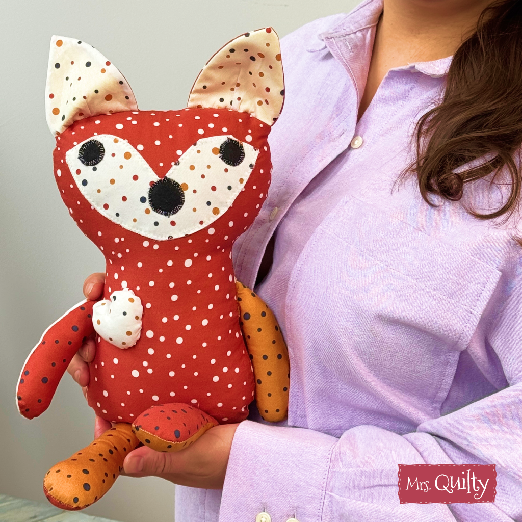 A person in a light purple shirt holds a red, polka-dotted fox plush made from the "Finley the Fox Downloadable PDF Quilt Pattern." The unique toy features mismatched arms and legs. The "Mrs. Quilty" logo appears at bottom right.