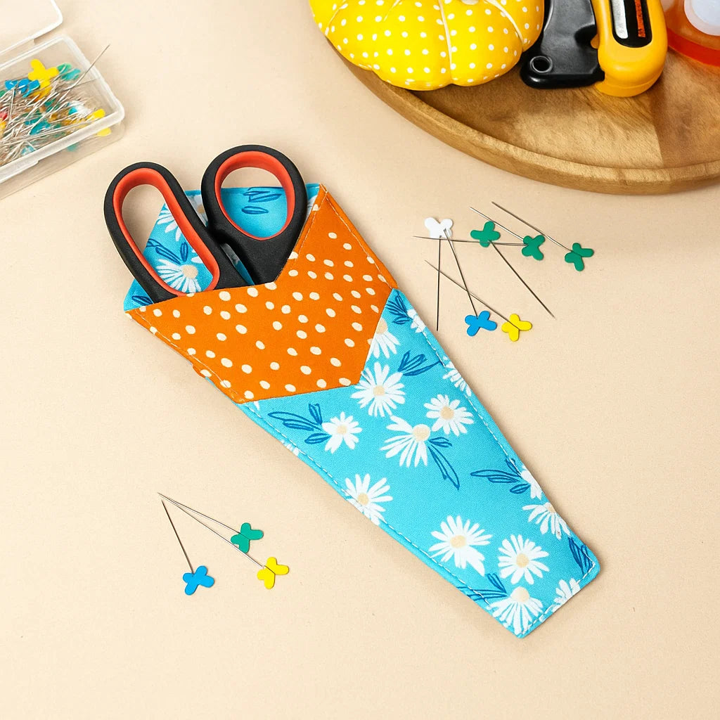 A pair of scissors in a blue and orange floral case made with the Functional Scissors Case Downloadable PDF Quilt Pattern, surrounded by sewing pins and tools on a beige surface.
