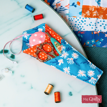 The Functional Scissors Case Downloadable PDF Quilt Pattern creates a floral fabric pouch that holds scissors, a seam ripper, and measuring tape—perfect for organizing quilting tools on your tabletop with fabrics and thread.