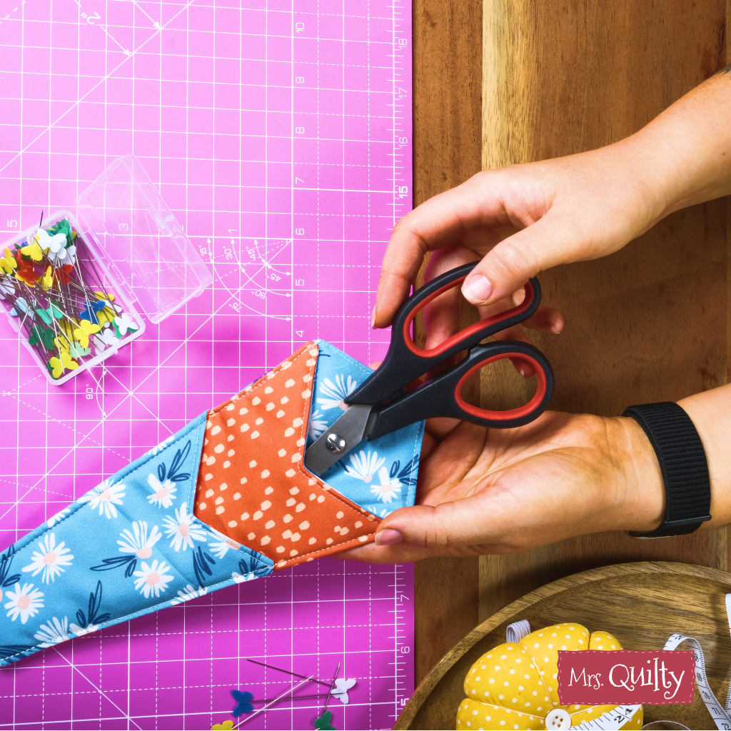 A person places scissors into a blue and orange floral case on a pink cutting mat with pins and fabric—made using the Functional Scissors Case Downloadable PDF Quilt Pattern. This handy accessory keeps your tools organized while quilting.