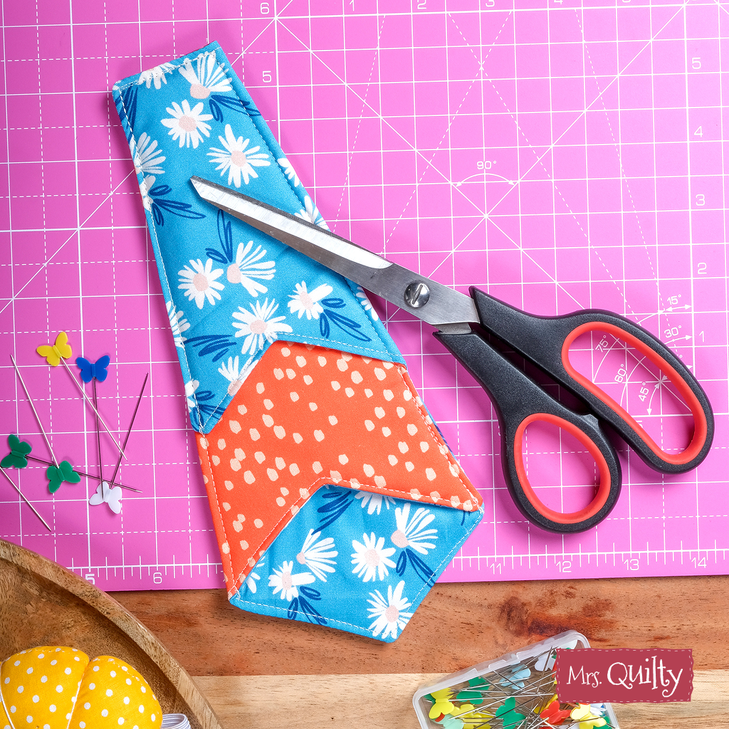 A pair of crafting scissors in a blue and orange floral case made from the Functional Scissors Case Downloadable PDF Quilt Pattern rests on a pink mat, surrounded by sewing pins, a pincushion, and fabric—an essential accessory for any quilting project.