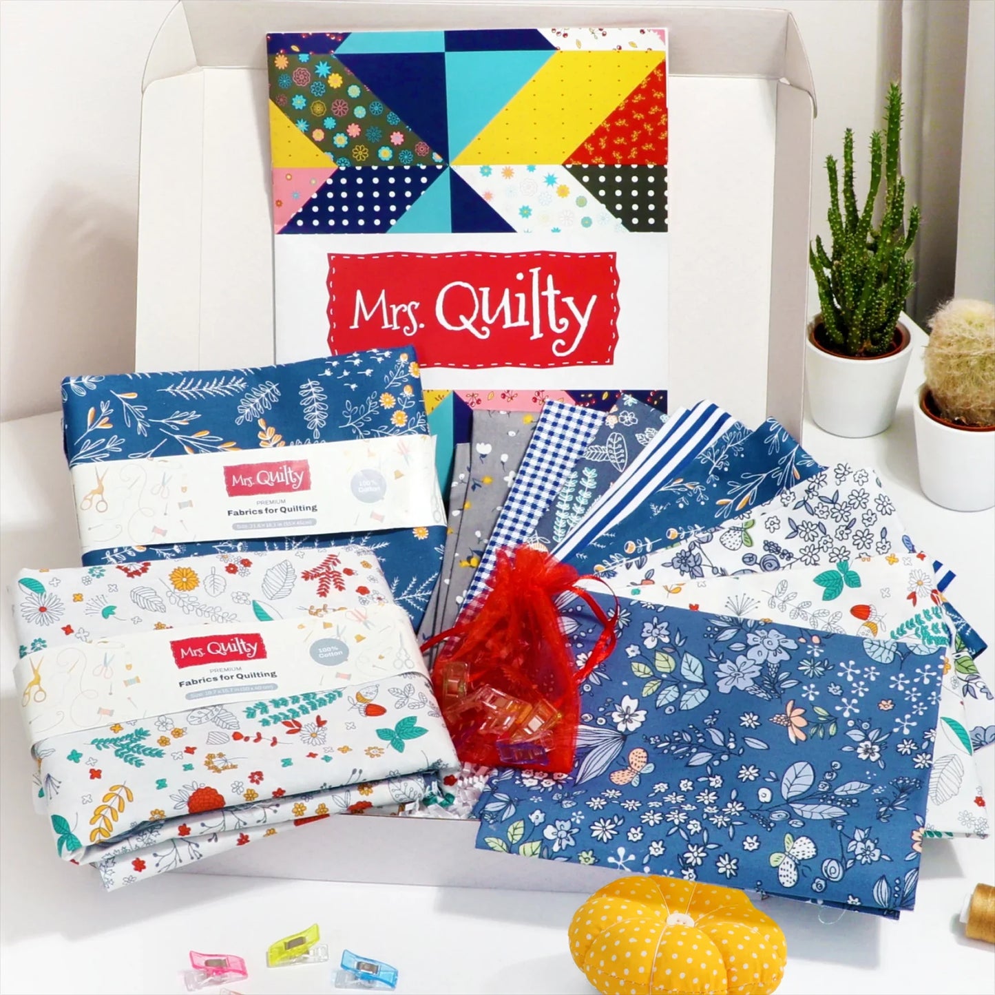 The Easter Bundle features a quilting fabric set in assorted blue and white patterns, accompanied by a Mrs. Quilty book, red pouch, sewing clips, colorful pin cushion, and a small cactus plant on a white surface.
