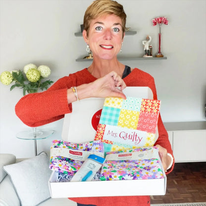 In a floral-themed living room, a person smiles while holding an Easter Bundle box containing colorful fabric squares, a pouch, and a skincare product.