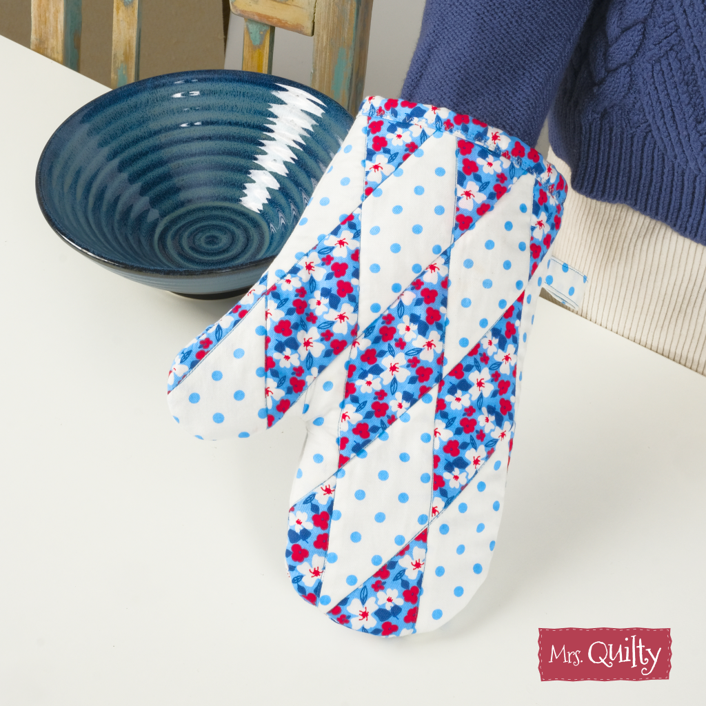 A person in a blue sweater holds the Oven Mitt Downloadable PDF Quilt Pattern, featuring floral, polka dot, and diamond designs—ideal for quilting enthusiasts—next to a dark blue bowl on a white table.