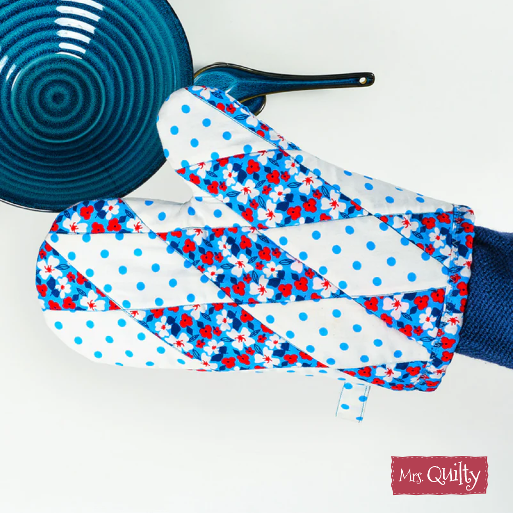 A hand wearing an oven mitt made with the "Oven Mitt Downloadable PDF Quilt Pattern" holds a blue ceramic bowl. The mitt features a blue, white, and red floral design. The Mrs. Quilty logo appears in the lower right corner.