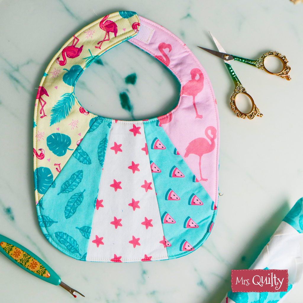 The Patchwork Baby Bib Downloadable PDF Quilt Pattern features flamingo, leaf, star, and watermelon designs—ideal for baby shower gifts or a quick DIY sewing project. Shown on marble with scissors and seam ripper; instant digital download.