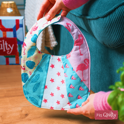 A person holds a Patchwork Baby Bib featuring flamingos, stars, watermelons, and leaves—made using the downloadable PDF quilt pattern from Mrs. Quilty. The logo appears in the bib’s bottom right corner, making it a great baby shower gift.