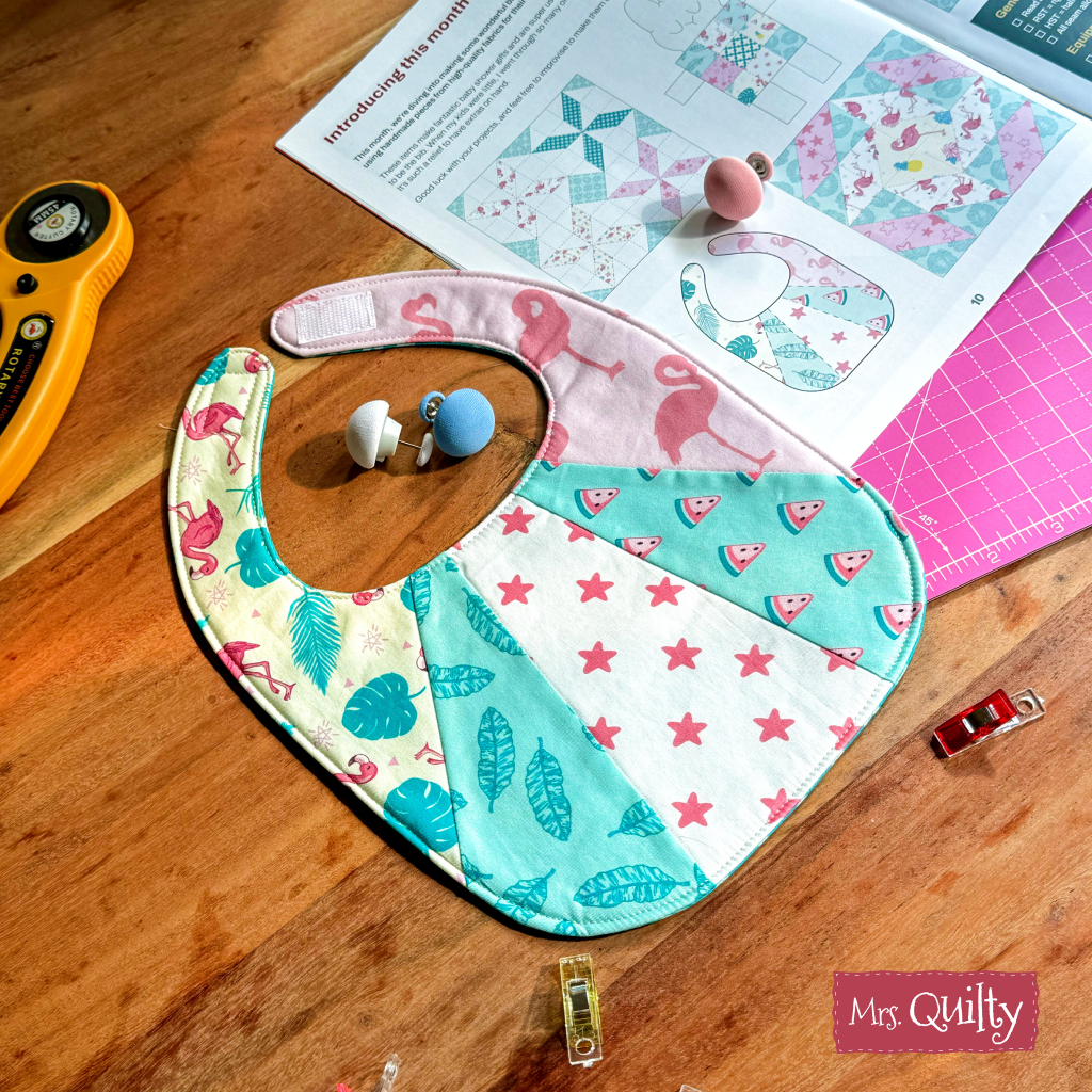 A Patchwork Baby Bib Downloadable PDF Quilt Pattern featuring tropical and star designs is displayed on a wooden table with sewing tools and a quilting magazine—ideal for crafting unique baby shower gifts.