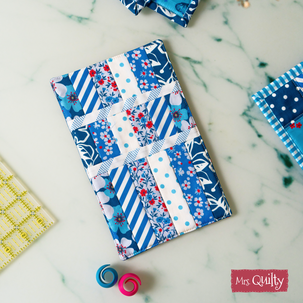 The Quilt-As-You-Go Mug Rug Downloadable PDF Quilt Pattern features blue, white, and floral patchwork strips—ideal for creative crafting and customizable gifts. Displayed on marble with sewing accessories.