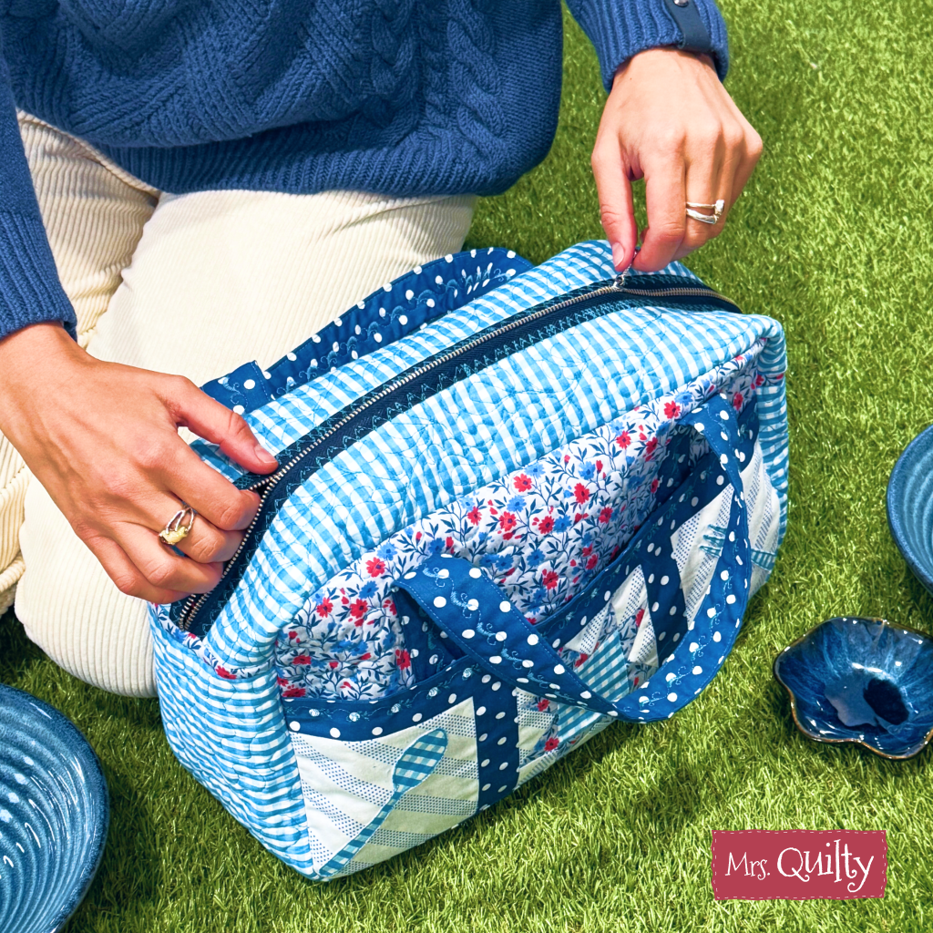 A person in light pants and a blue sweater opens a lunch bag on the grass near ceramic dishes, featuring the "Quilter’s Lunch Bag Downloadable PDF Quilt Pattern" and showcasing creative quilting techniques.