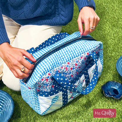 A person in light pants and a blue sweater opens a lunch bag on the grass near ceramic dishes, featuring the "Quilter’s Lunch Bag Downloadable PDF Quilt Pattern" and showcasing creative quilting techniques.